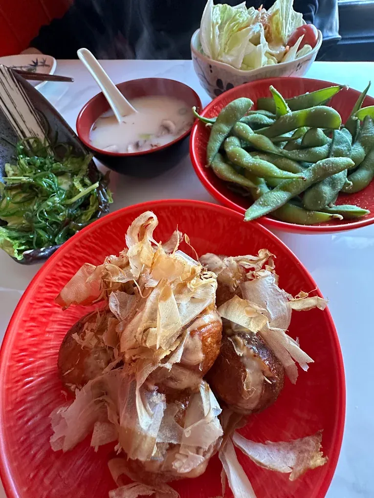 Takoyaki topped with bonito flakes on a red plate, with edamame and soup behind it.